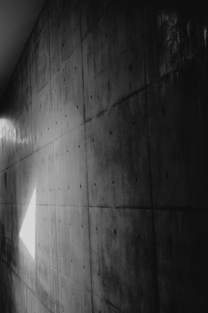 Muro de concreto en penumbra con un haz de luz cenital entrando por un vano, formando un triángulo de luz nítido en la oscuridad